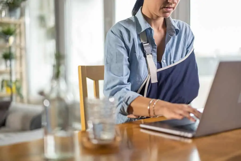 A woman, with her arm in a sling, types on her computer.