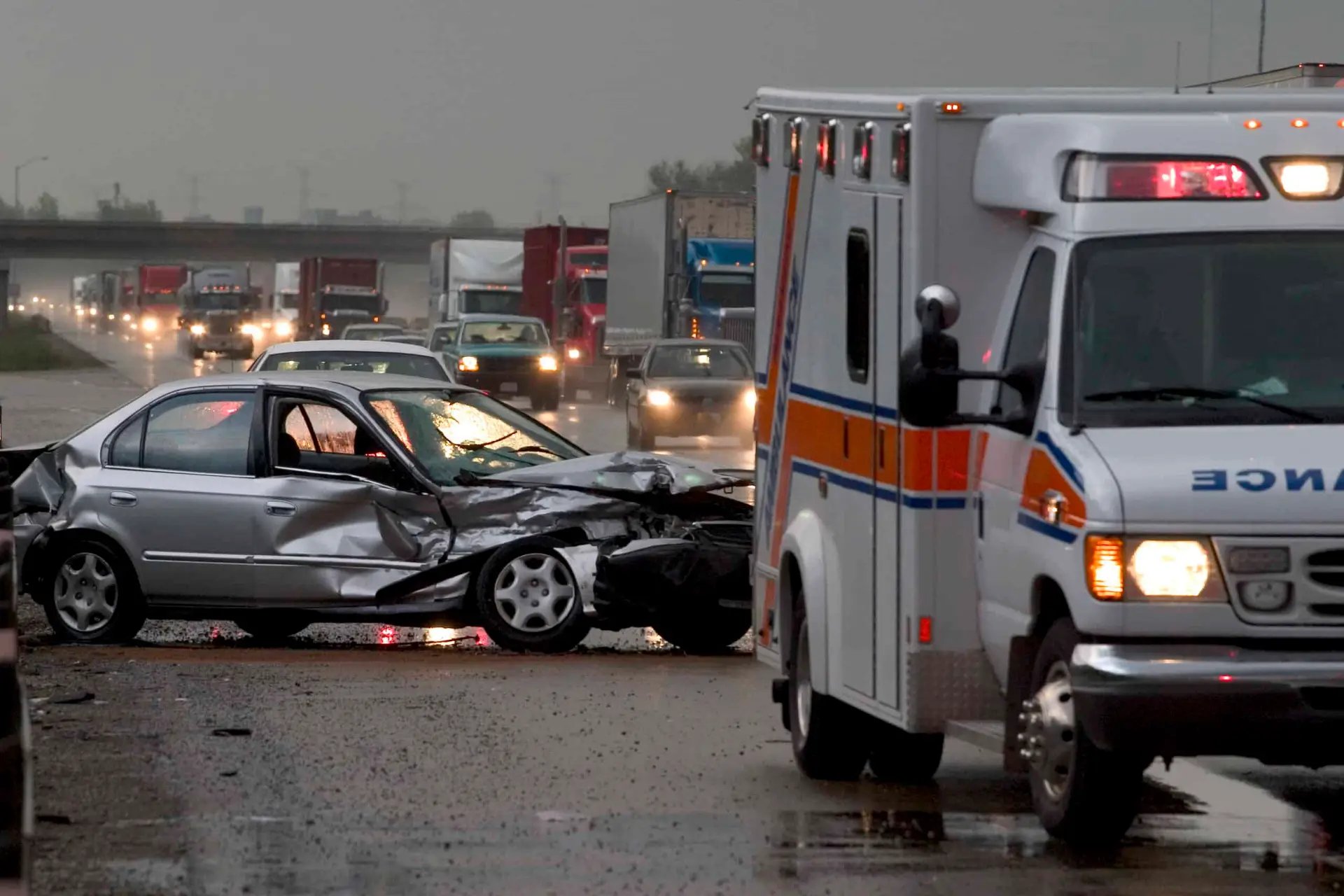 An ambulance arrives at the scene of a car accident on the side of a busy highway on a rainy afternoon.