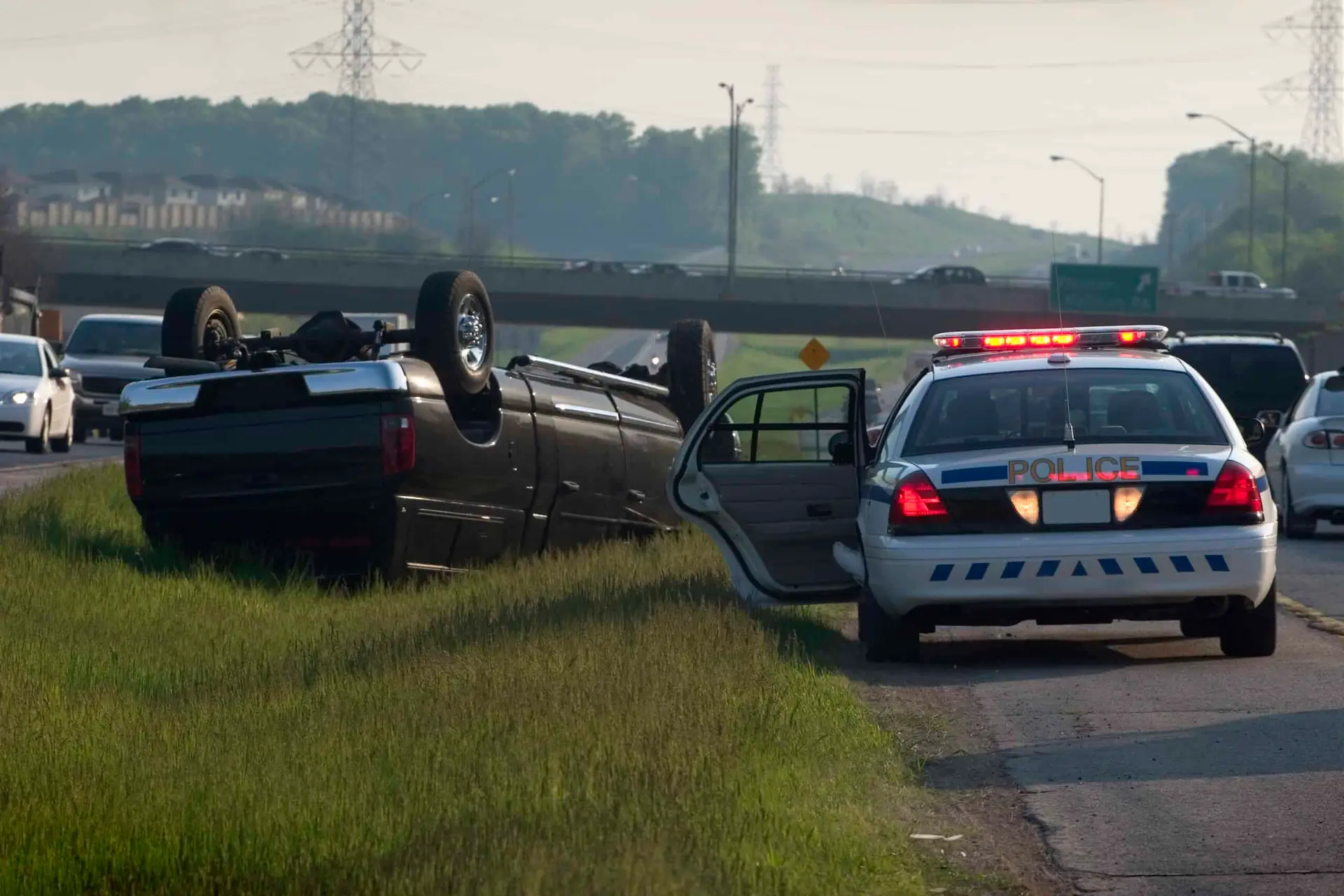 A large SUV rolled over in the middle of a busy median off of the highway. A police vehicle is on the scene. 