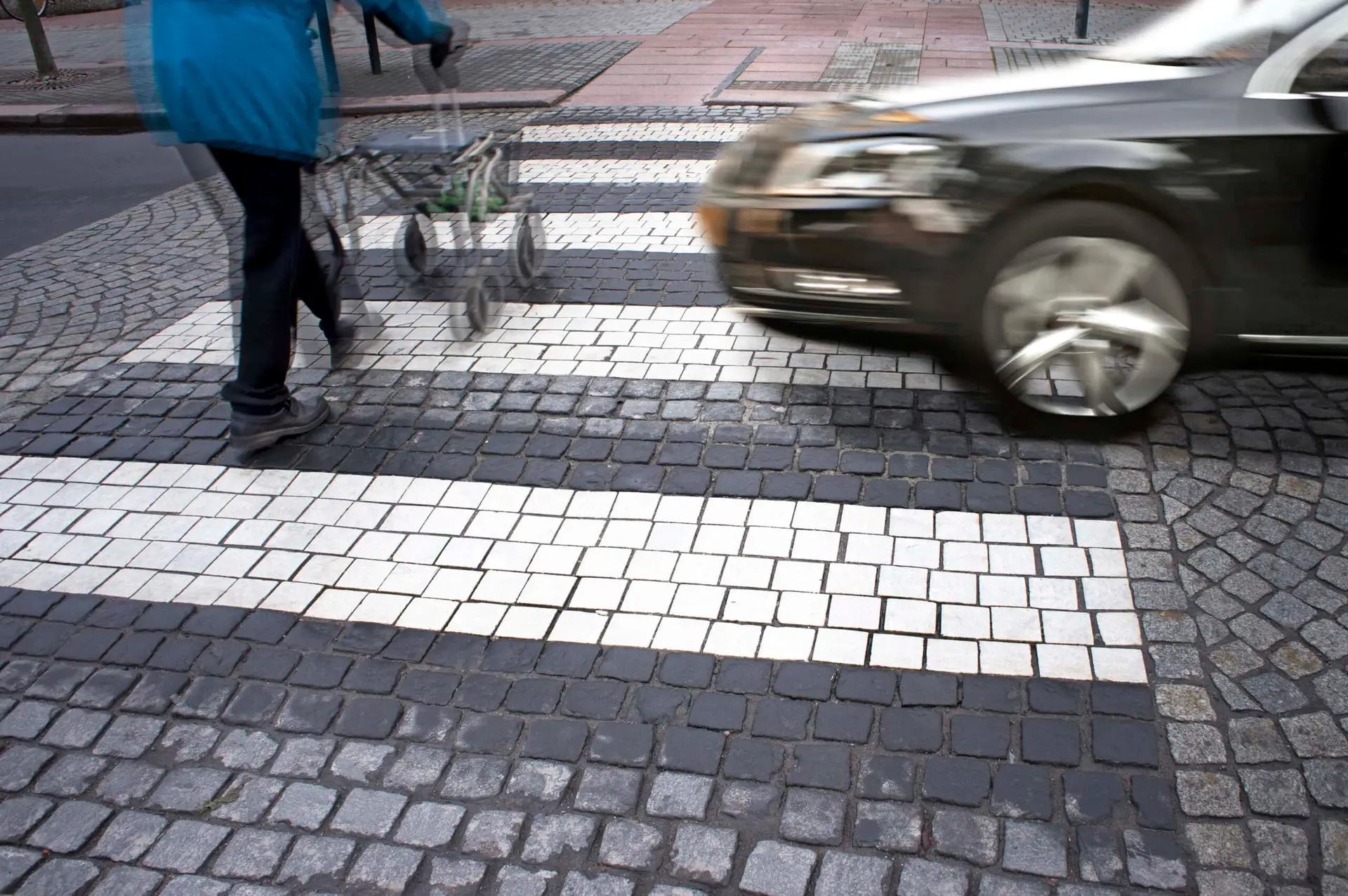 A person using a walker crossing at a crosswalk is about to get hit by an approaching black car.