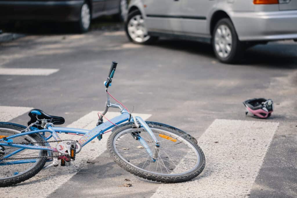 A damaged bicycle rests in the middle of a crosswalk with a bike helmet close by after a car and bicycle collide.