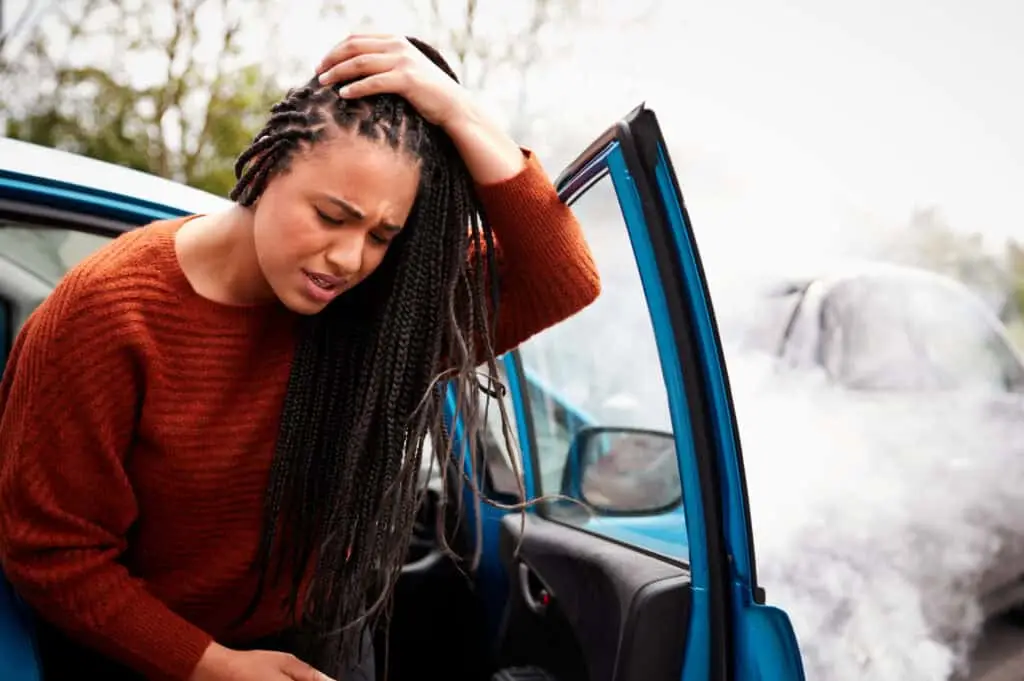 A woman exits a car after an accident holding her head and wincing in pain. 