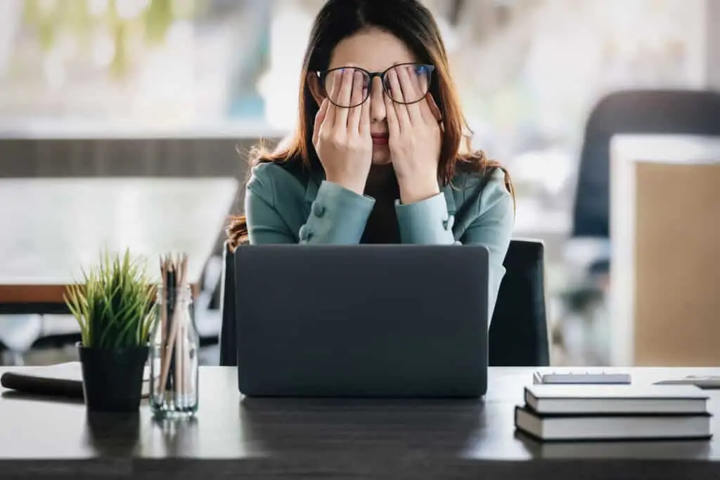 A woman rubs her eyes and rests her head in her hands while sitting at a work desk, showing signs of mental fatigue