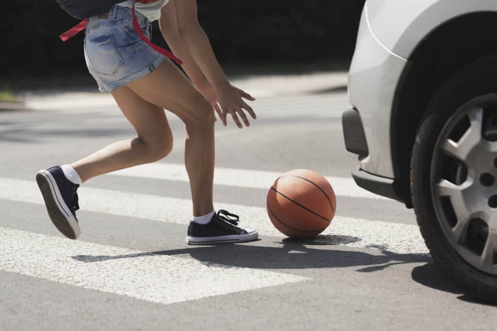 A young girl stoops down to pick up a basketball in an intersection crosswalk with a white car approaching from the side.