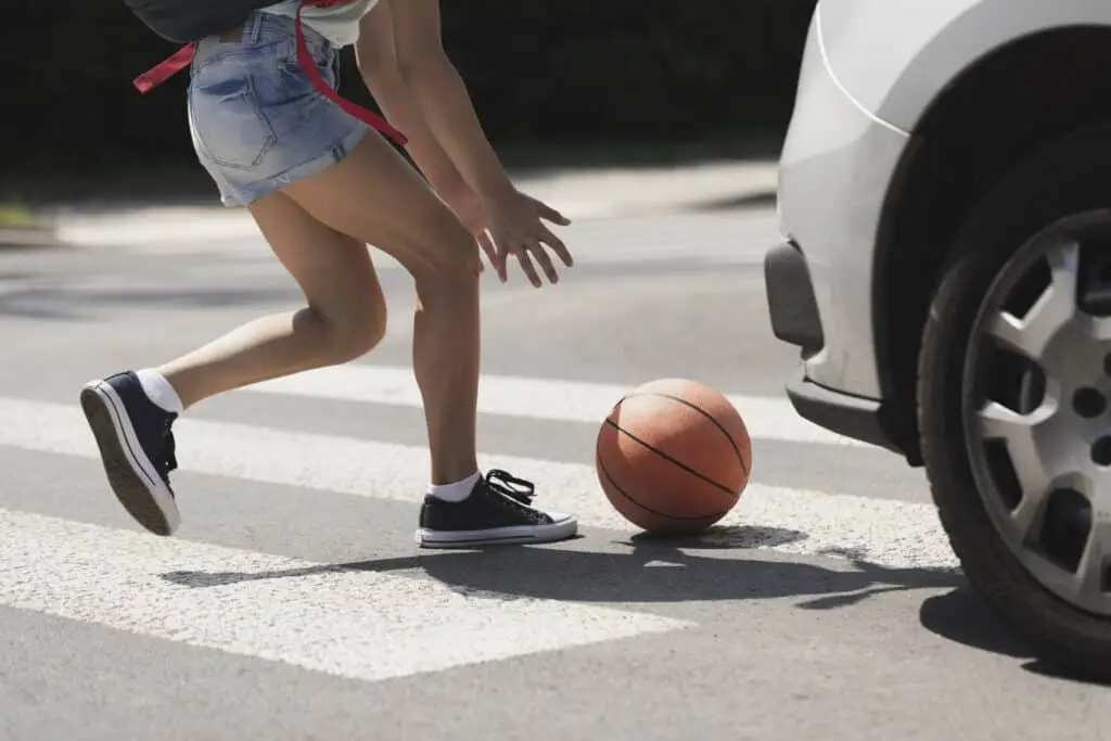 A young girl stoops down to pick up a basketball in an intersection crosswalk with a white car approaching from the side.
