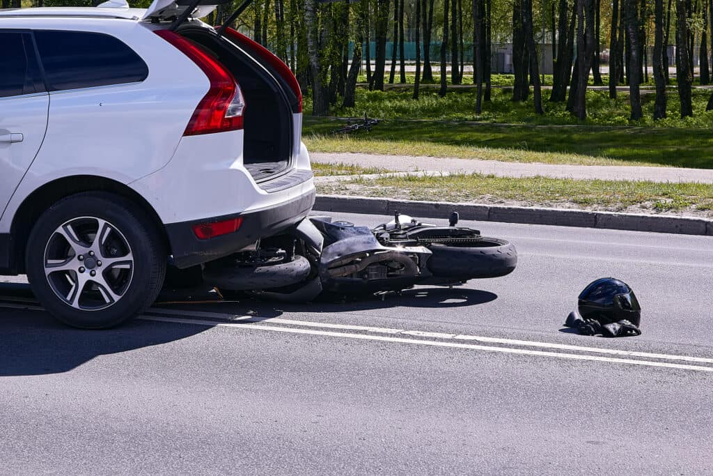 An accident scene where a minivan has collided with a motorcycle. The minivan is on top of the motorcycle and the motorcycle riders helmet and gloves are on the ground in the middle of the road.