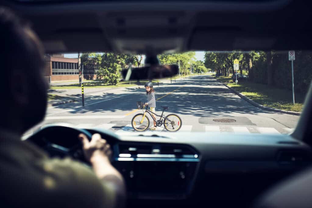 A person driving a car approaches a child pedestrian in the crosswalk walking their bicycle across the road.