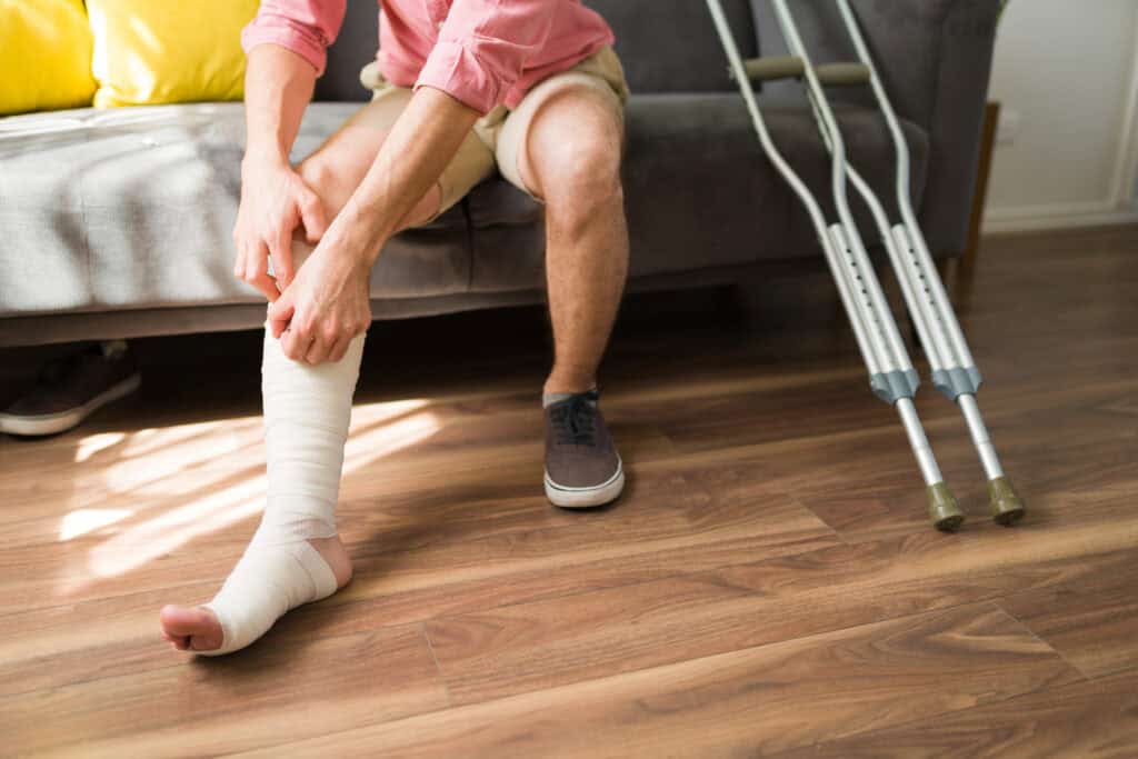 A man adjusts the wrappings around his leg while he sits on a couch, with crutches propped up against it.