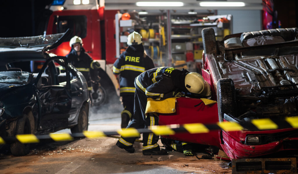 First responders at the scene of a fatal car accident on the highway at night. A red car is flipped over on its hood and a firefighter is surveying the interior of the vehicle.