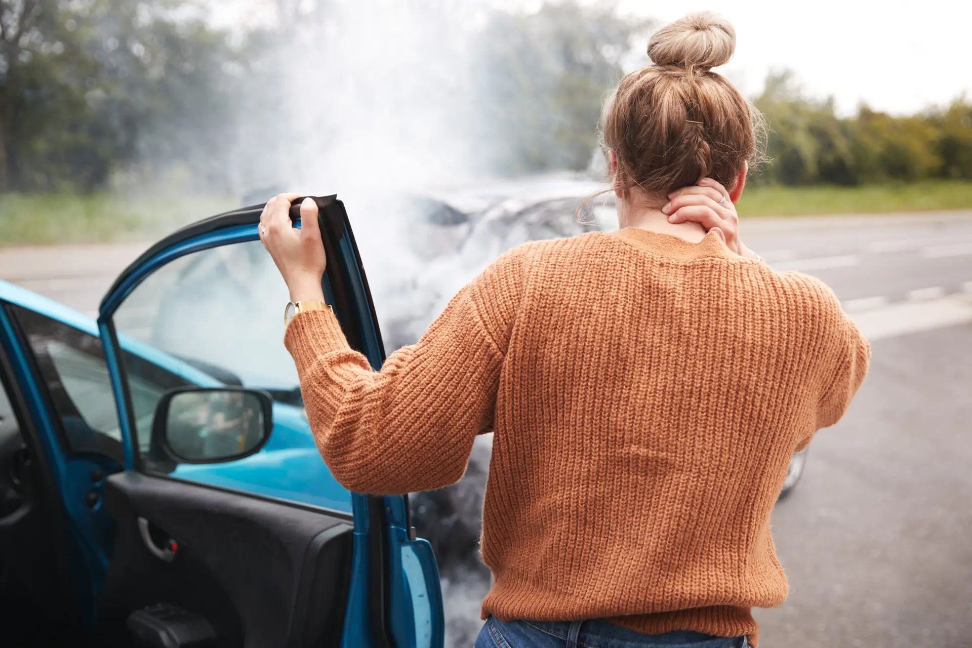 Woman at a car accident touching her neck beside a damaged, smoking vehicle, suggesting possible injury