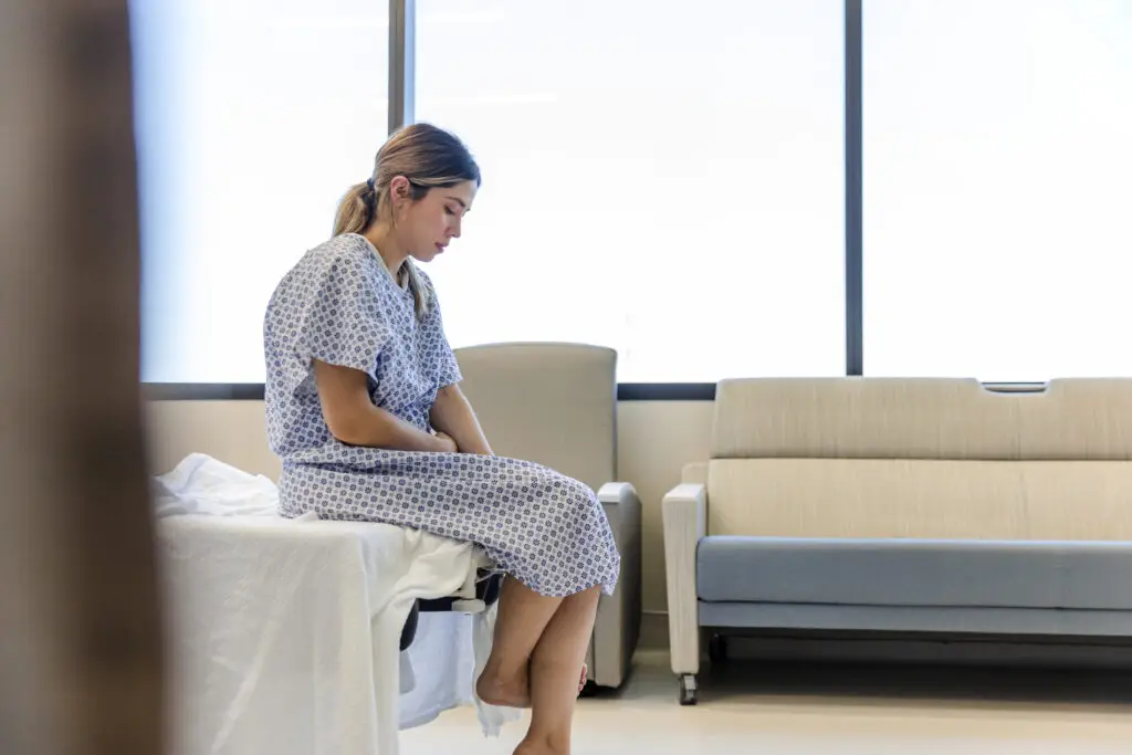 A woman looks depressed while she sits at the end of her hospital bed.