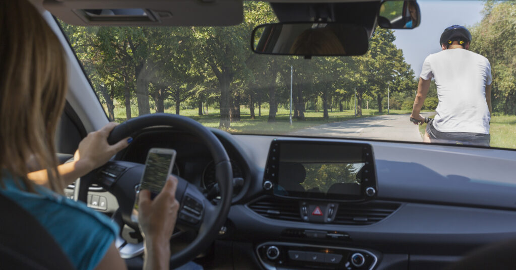 A woman is texting and driving, not paying attention to the road. She is approaching a cyclist from behind. 