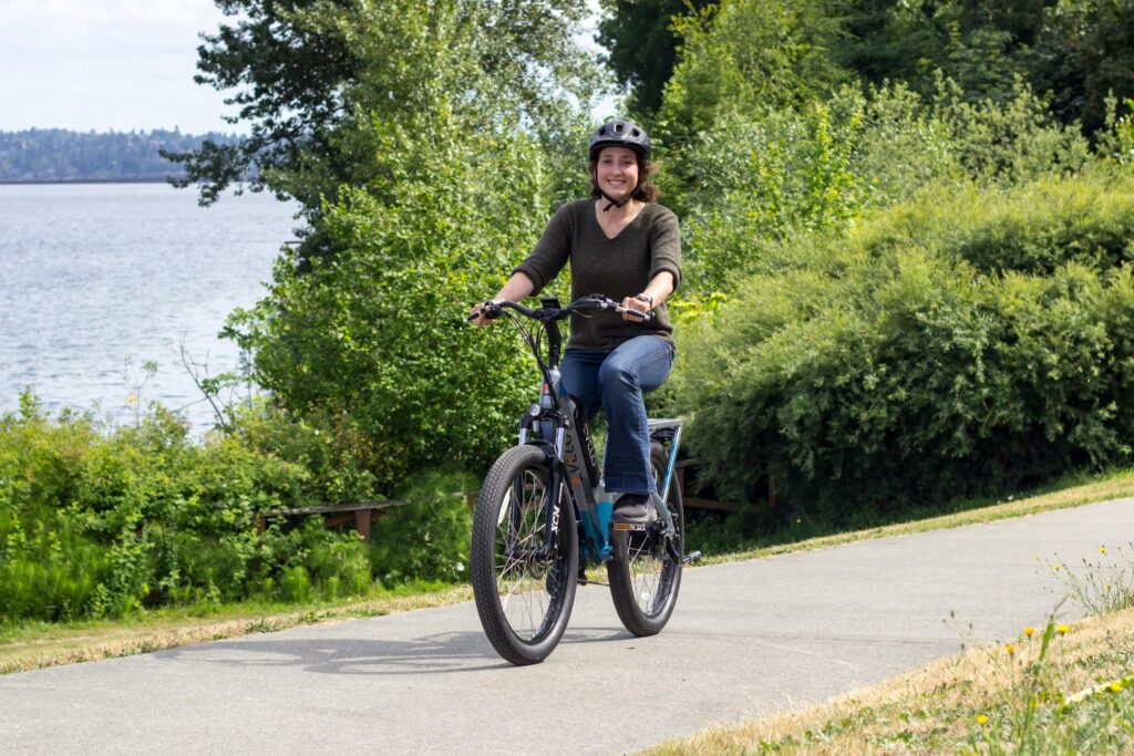 A woman rides an electric bicycle along a lakeside path on a sunny day. 